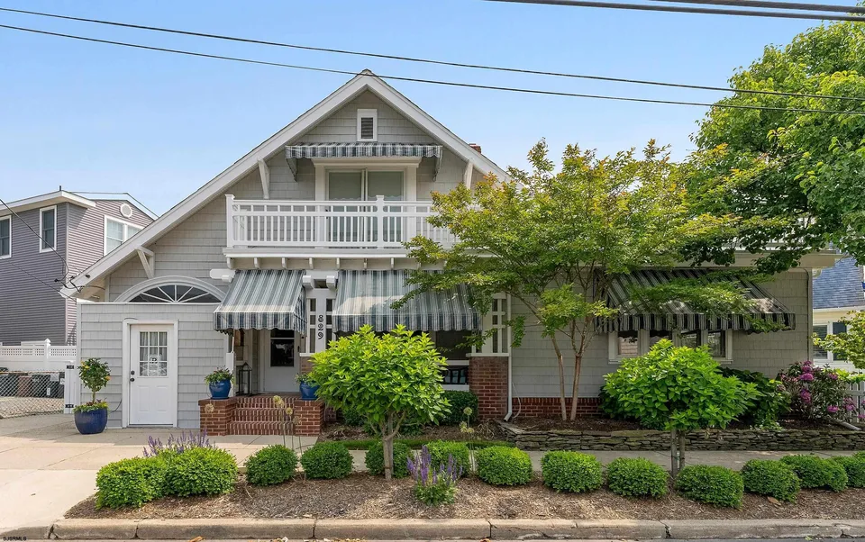 We see the front of a grey house adorned by trees and plants in Ocean City, NJ against a clear blue sky.